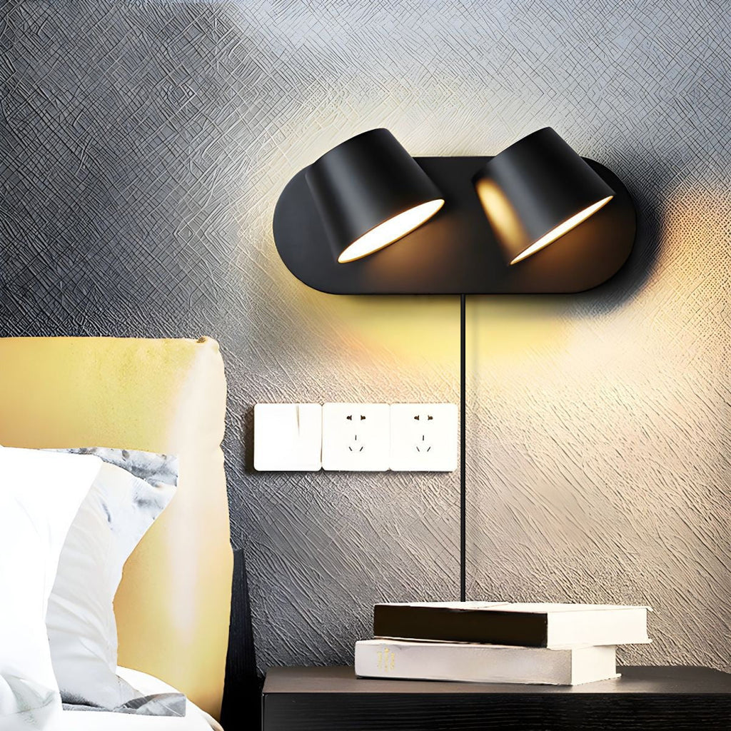 Two-light black Clérachic wall light glowing warm on a dark textured wall above a bedside table, with a row of sockets and a stack of books.