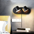 Two-light black Clérachic wall light glowing warm on a dark textured wall above a bedside table, with a row of sockets and a stack of books.