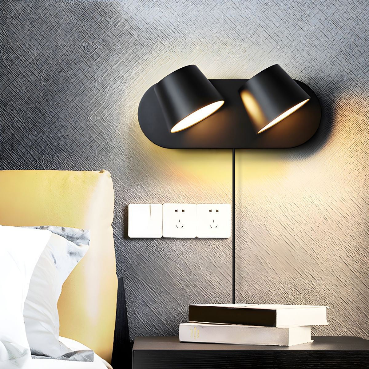 Two-light black Clérachic wall light glowing warm on a dark textured wall above a bedside table, with a row of sockets and a stack of books.