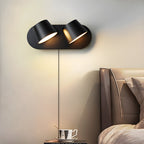 Black Clérachic wall light with two tilted shades above a bedside, its cord running down a cream wall beside a leather headboard and a cup.