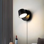 Single-light black Clérachic wall light turned to the side on a cream wall, beside a vertical slatted panel and a wine bottle.