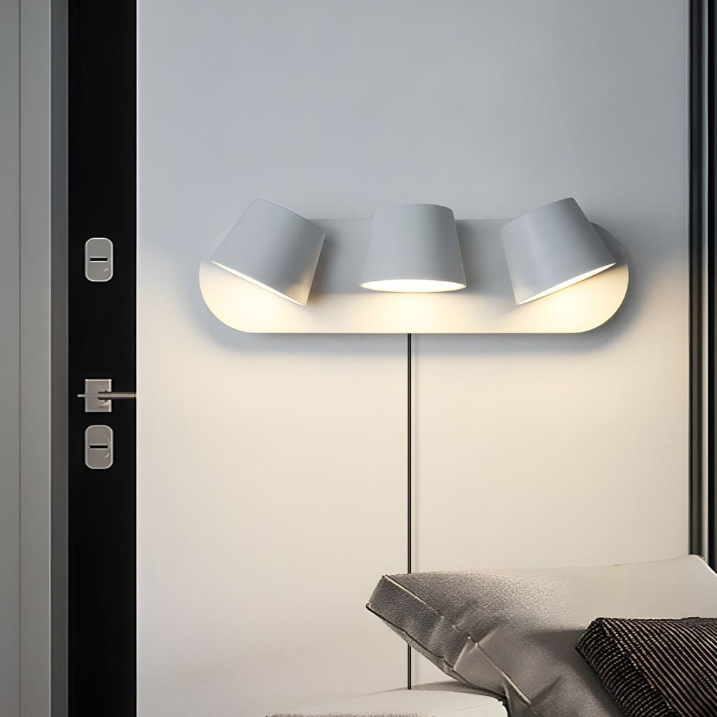 Three-light white Clérachic wall light on a grey wall beside a black door, the shades angled in different directions above a bed.