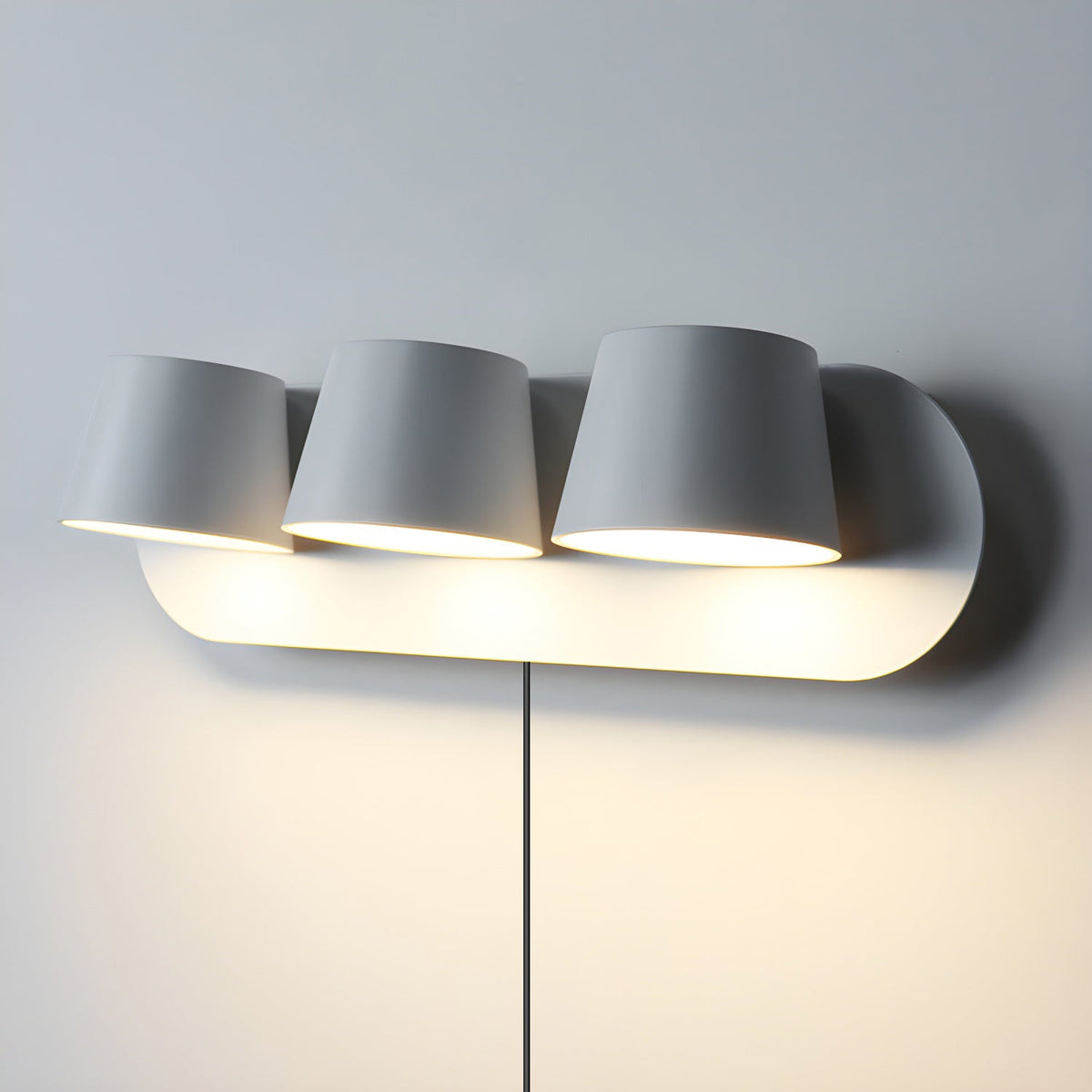 Three-light white Clérachic wall light on a pale wall, the shades lit warm on a long oval plate with its cord hanging straight down.