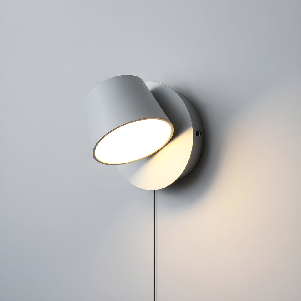 Single-light white Clérachic wall light with its round head tilted, glowing warm on a pale grey wall with the cord hanging below.