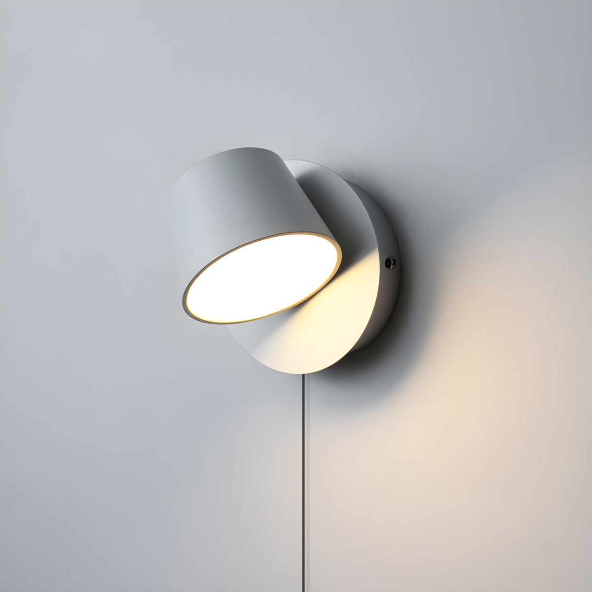 Single-light white Clérachic wall light with its round head tilted, glowing warm on a pale grey wall with the cord hanging below.