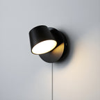 Single-light black Clérachic wall light with the head turned to face the viewer, lit warm on a pale grey wall.