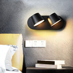 Black two-light Clérachic wall light on a dark textured wall, glowing warm above a pale socket plate and a stack of books on a nightstand.