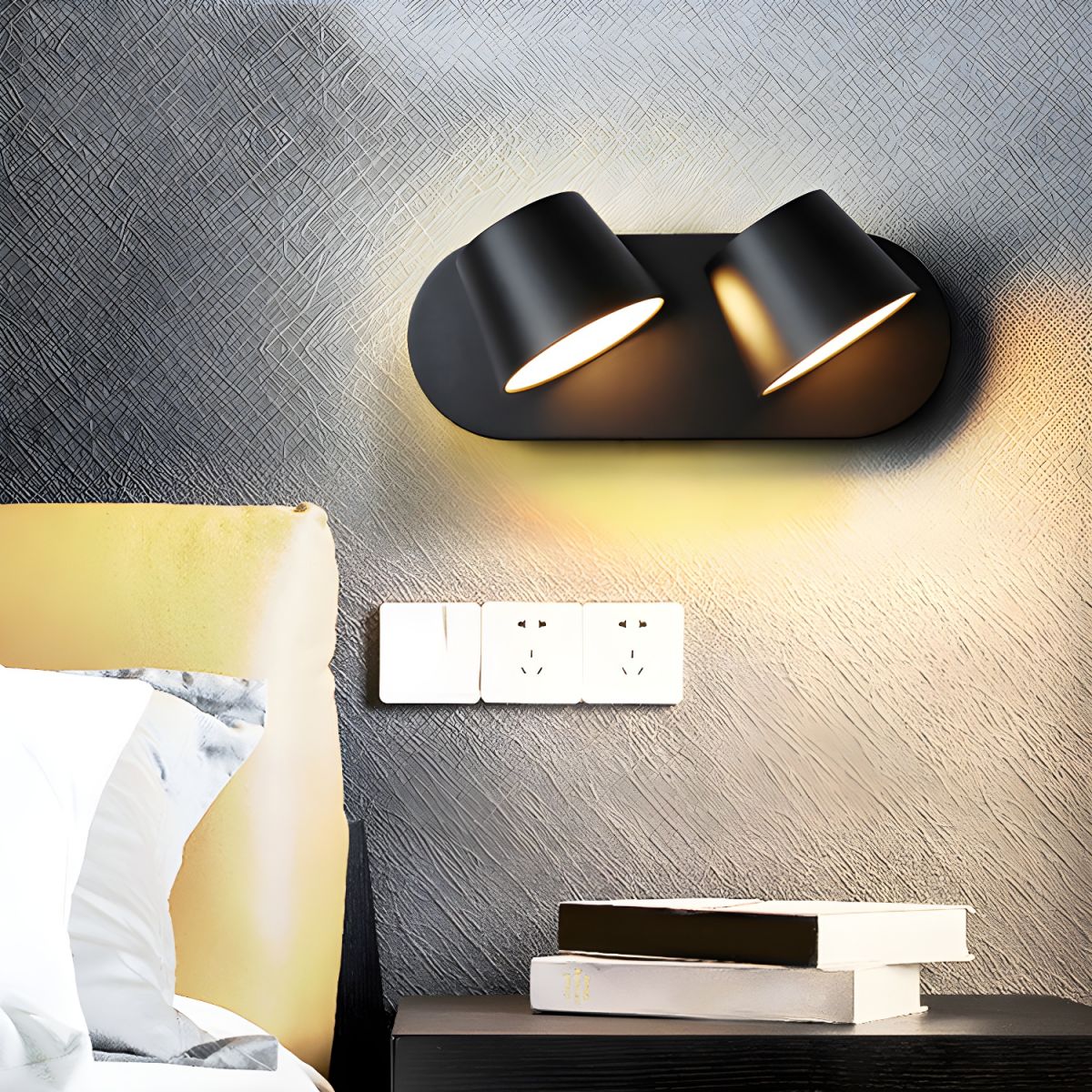 Black two-light Clérachic wall light on a dark textured wall, glowing warm above a pale socket plate and a stack of books on a nightstand.