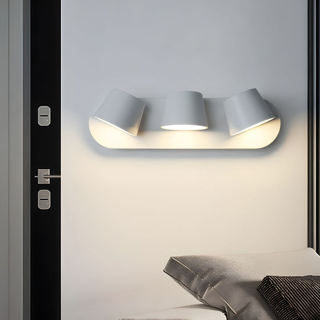 White Clérachic wall light with three shades above a bed corner, fixed beside a black door frame and lit warm on the wall.
