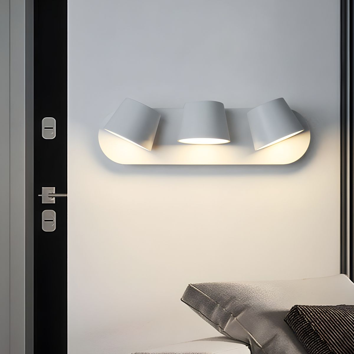 White Clérachic wall light with three shades above a bed corner, fixed beside a black door frame and lit warm on the wall.