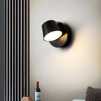Black single-light Clérachic wall light on a cream wall beside a slatted panel, with a wine bottle and a woven cushion below.
