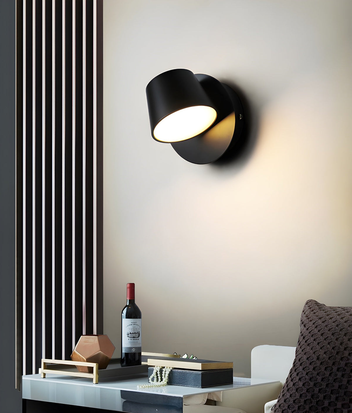 Black single-light Clérachic wall light on a cream wall beside a slatted panel, above a tray with a wine bottle and small ornaments.