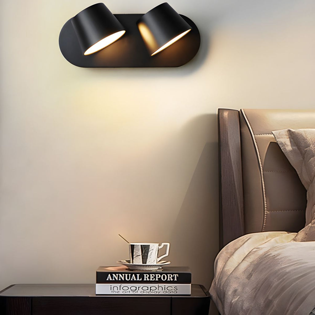Two-light black Clérachic wall light above a dark bedside table, with a cup and saucer on a stack of books and a headboard to the right.