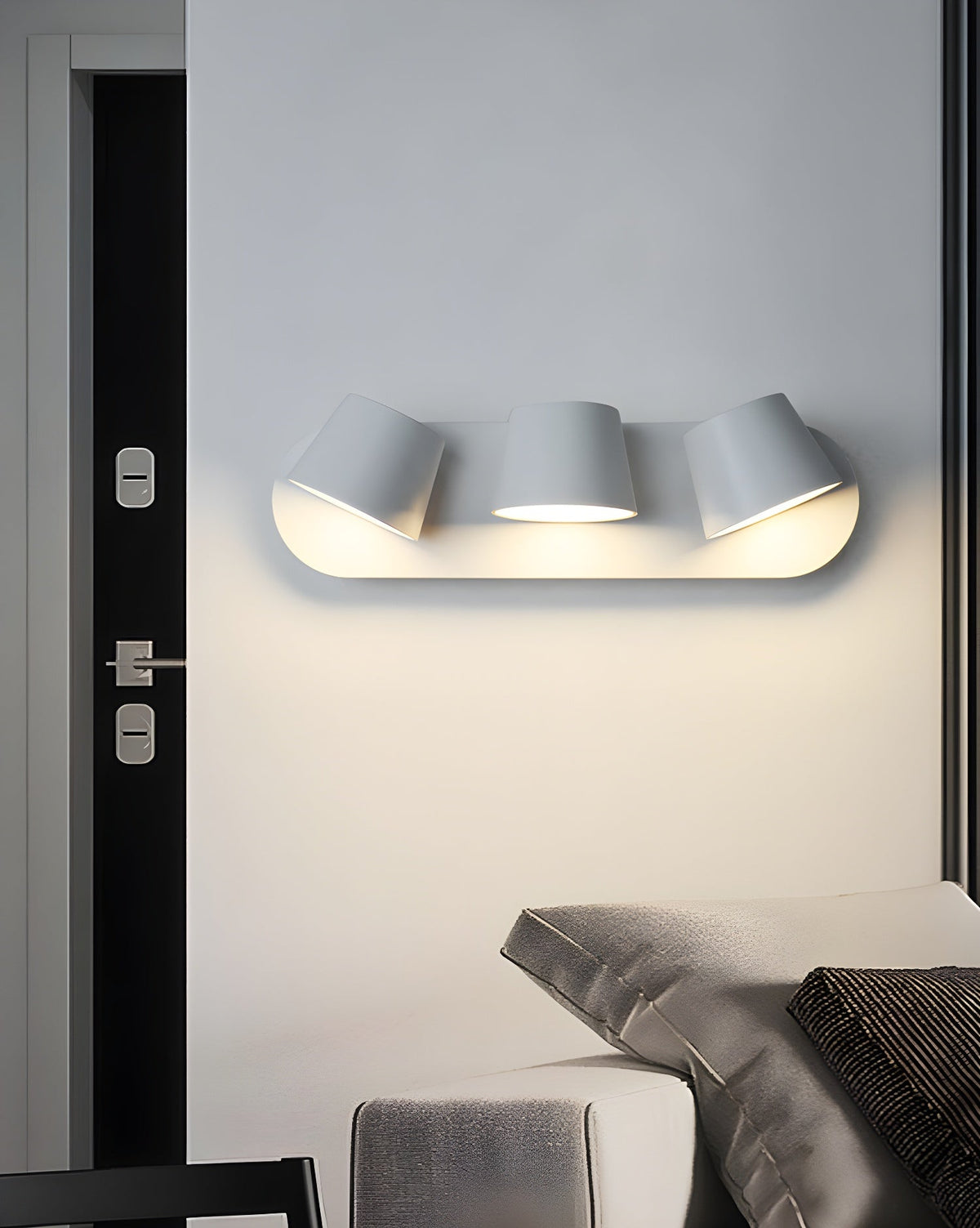Three-light white Clérachic wall light on a pale wall beside a black door with chrome handles, with a bed's grey cushions below.