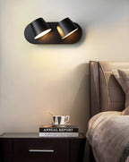 Black Clérachic wall light with two shades and a pale cream wall behind, with a dark nightstand and a bed's headboard below.