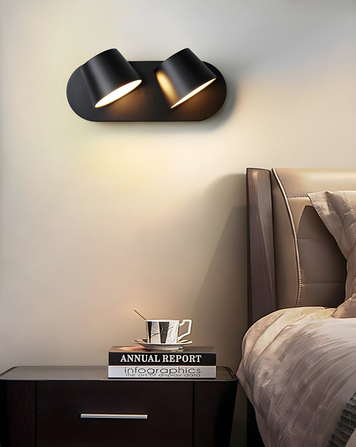 Black Clérachic wall light with two shades and a pale cream wall behind, with a dark nightstand and a bed's headboard below.
