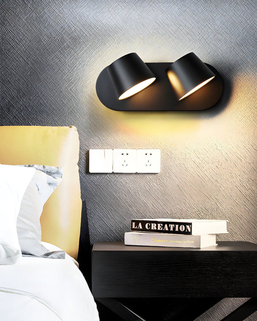 Black two-light Clérachic wall light on a dark textured wall above a nightstand, with sockets and books beside a cushioned headboard.