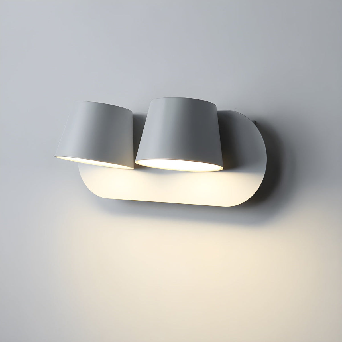 Close view of the two-light white Clérachic wall light, its tapered shades lit and washing the grey wall beneath with warm light.