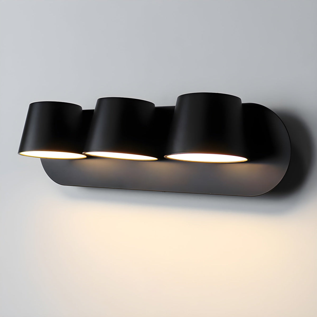 Close view of the three-light black Clérachic wall light, three tapered shades lit warm on a grey wall above a soft glow.