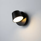 Close view of the single-light black Clérachic wall light on a pale grey wall, its round head tilted and lit with a warm glow.
