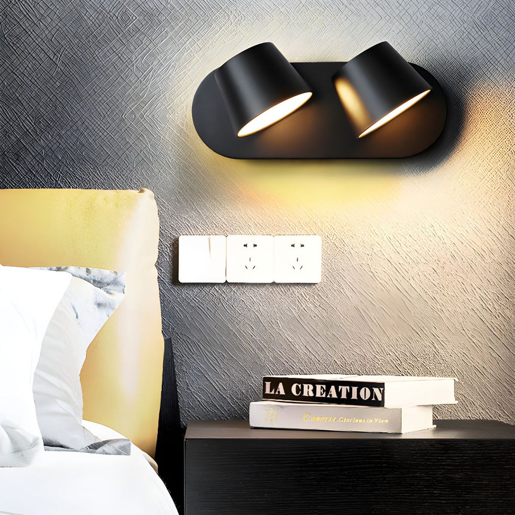 Black two-light Clérachic wall light on a dark textured wall above a nightstand, with sockets, two books and a cushioned headboard.