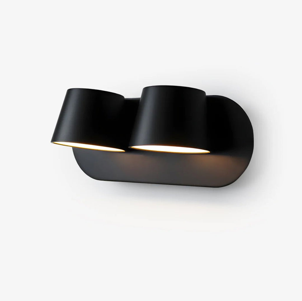 Black Clérachic wall light with two cylinder shades on a rounded oval plate, angled slightly down on a plain white backdrop.