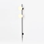 A Clérachic plug-in wall light with two frosted white globes on a slim black rod, with the cord trailing down a plain white wall.