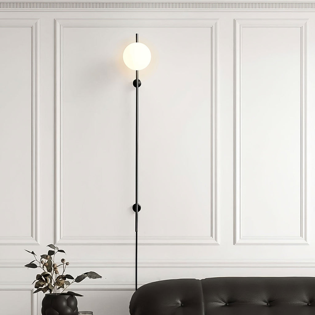 A Clérachic plug-in wall light with a single glowing globe on a thin black rod, against white wall panelling above a black leather sofa.