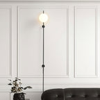 A Clérachic plug-in wall light with a single glowing globe on a thin black rod, against white wall panelling above a black leather sofa.