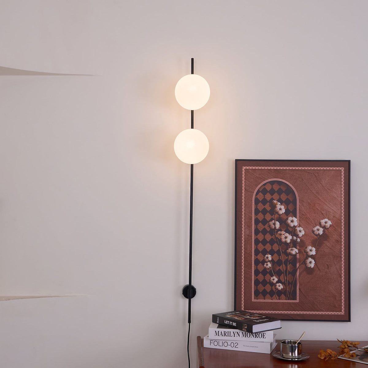 A Clérachic wall light with two softly glowing globes beside a framed rust-red print, with the black cord running down to a wooden console.