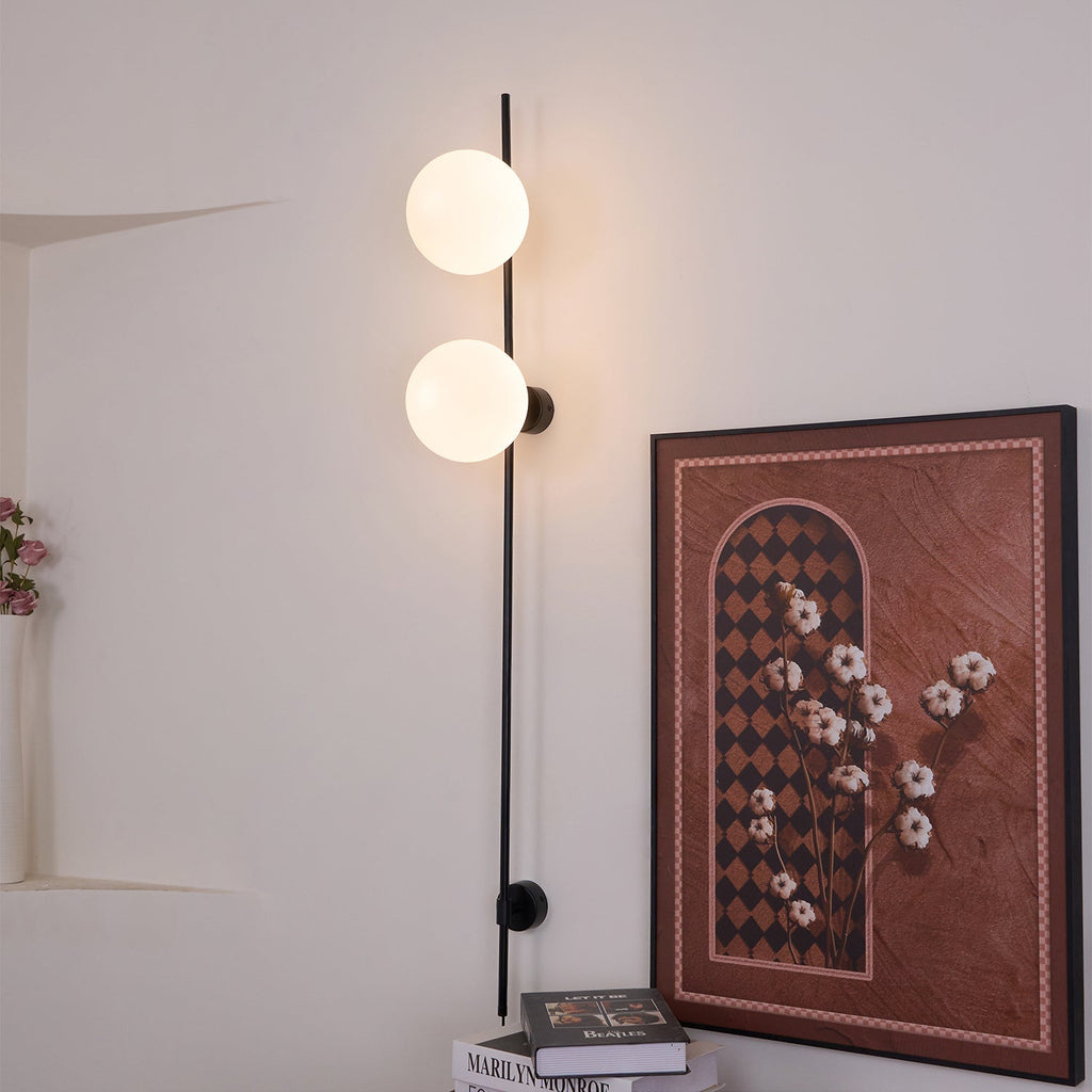 The Clérachic wall light lit, with two larger globes on its black rod, next to a framed floral print and a stack of books.