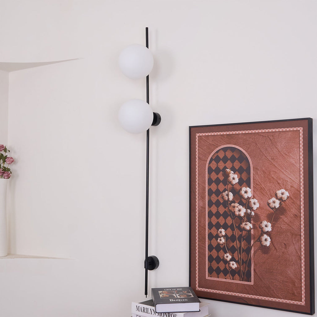 Unlit, this Clérachic wall light shows two milky white globes on a slim black rod, beside a large framed print and books on a shelf.