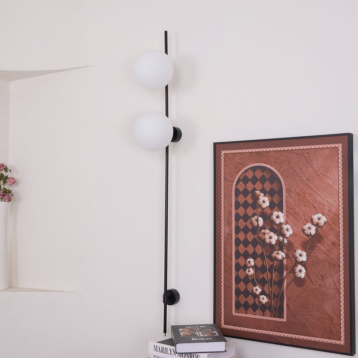Unlit, this Clérachic wall light shows two milky white globes on a slim black rod, beside a large framed print and books on a shelf.