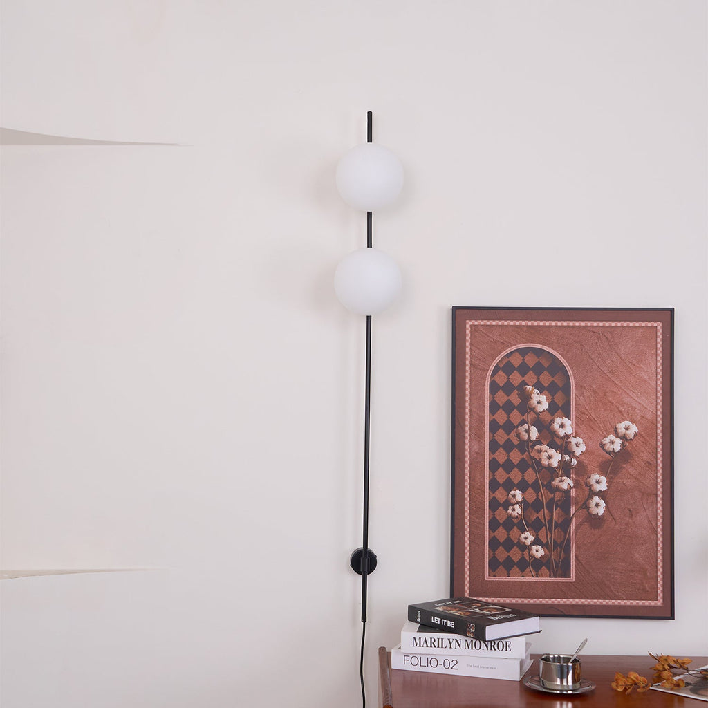 Both globes unlit on this Clérachic black wall light, hung on a white wall beside a framed print with a book stack below.
