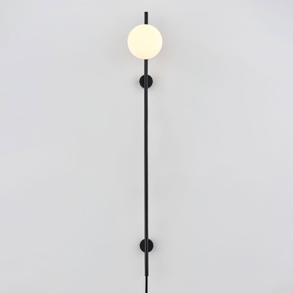 A Clérachic plug-in wall light with one white globe at the top of a long black rod, held by two small wall mounts on a pale wall.
