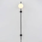A Clérachic plug-in wall light with one white globe at the top of a long black rod, held by two small wall mounts on a pale wall.