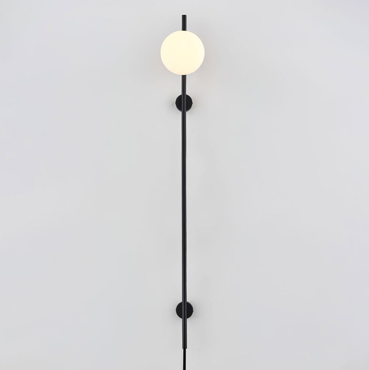 A Clérachic plug-in wall light with one white globe at the top of a long black rod, held by two small wall mounts on a pale wall.