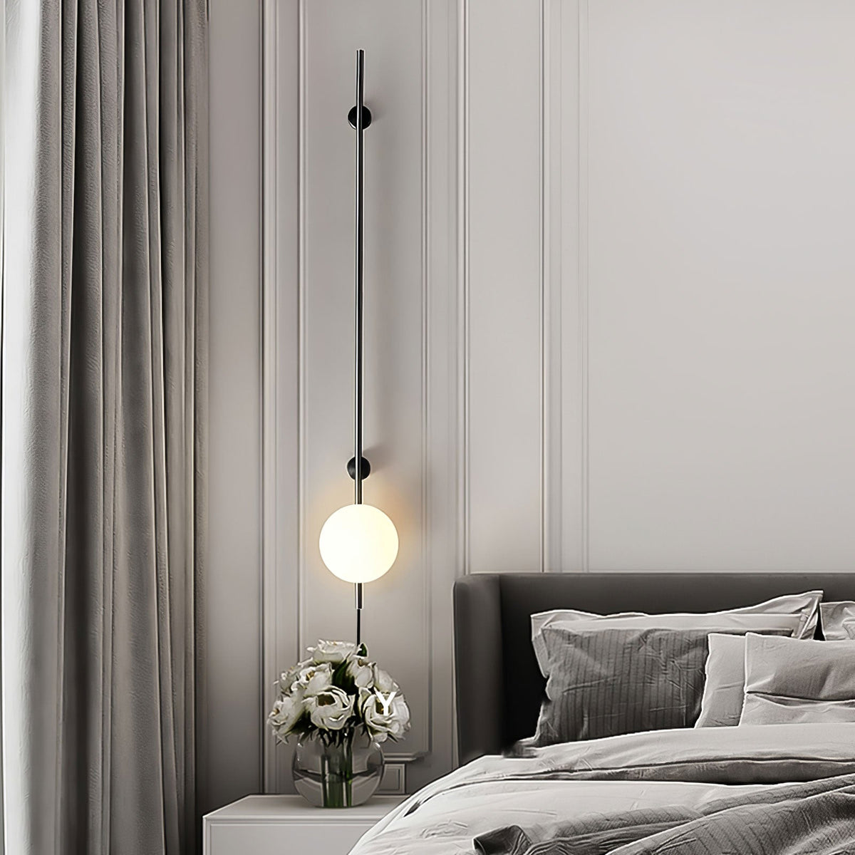 A Clérachic wall light with a lit globe low on a long black rod, beside a bedside table with white flowers and a grey bed.