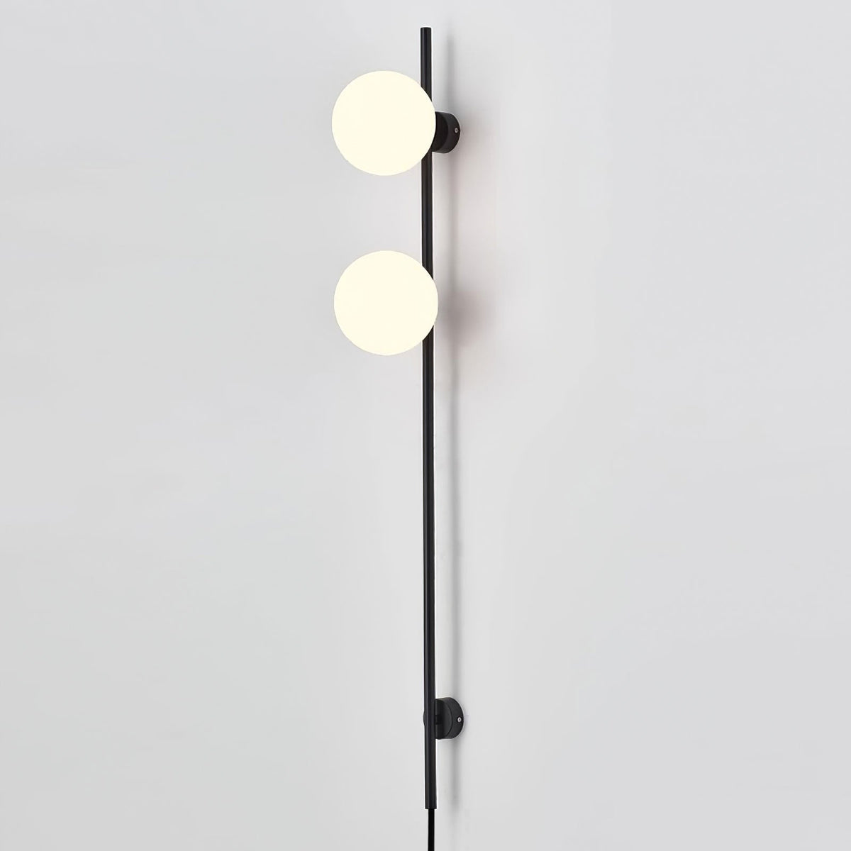 Close view of two glowing globes on this Clérachic slim black wall light, against a pale grey wall with the cord at the base.