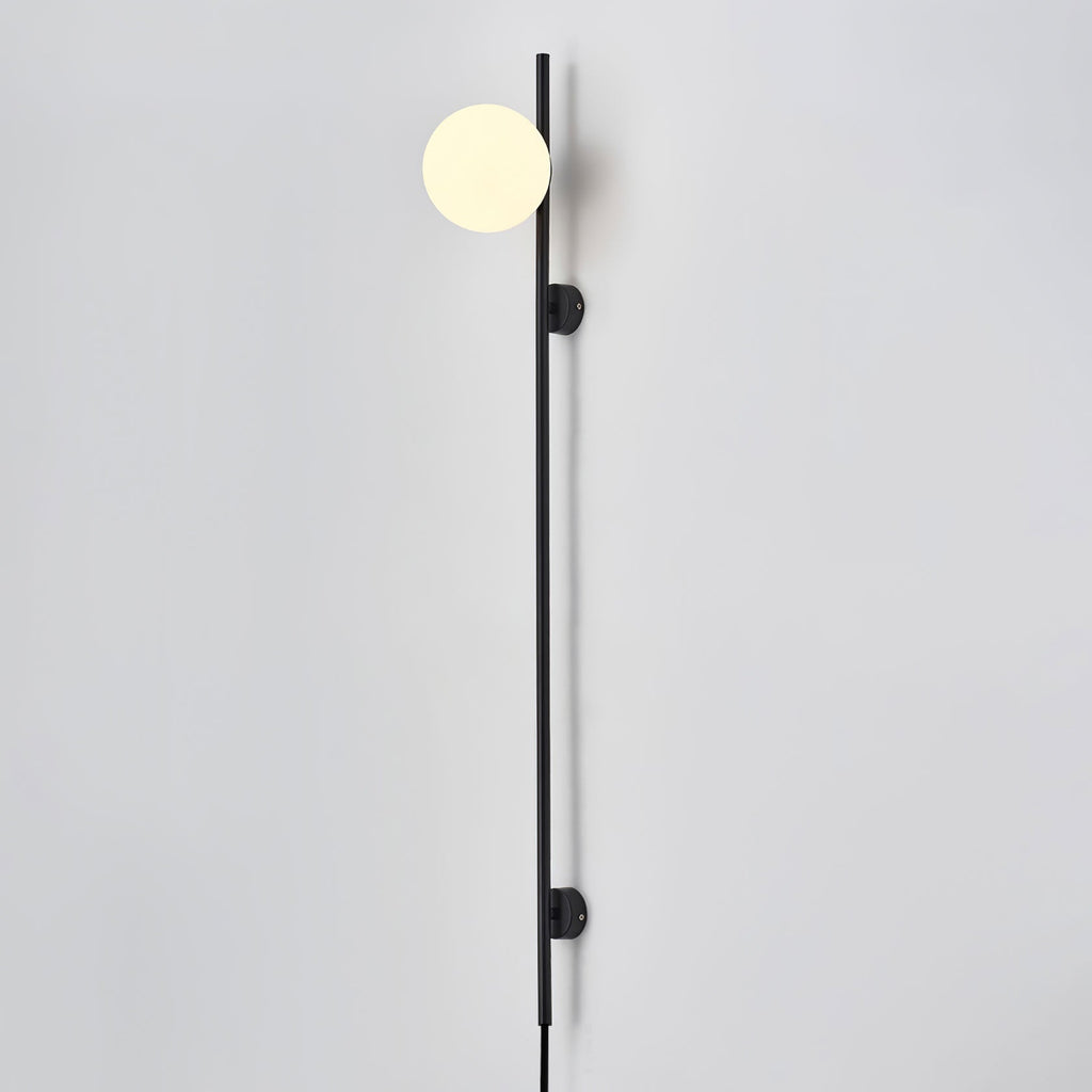 A Clérachic black wall light with one warm globe at the top of its rod, mounted on a pale wall with the cord hanging below.