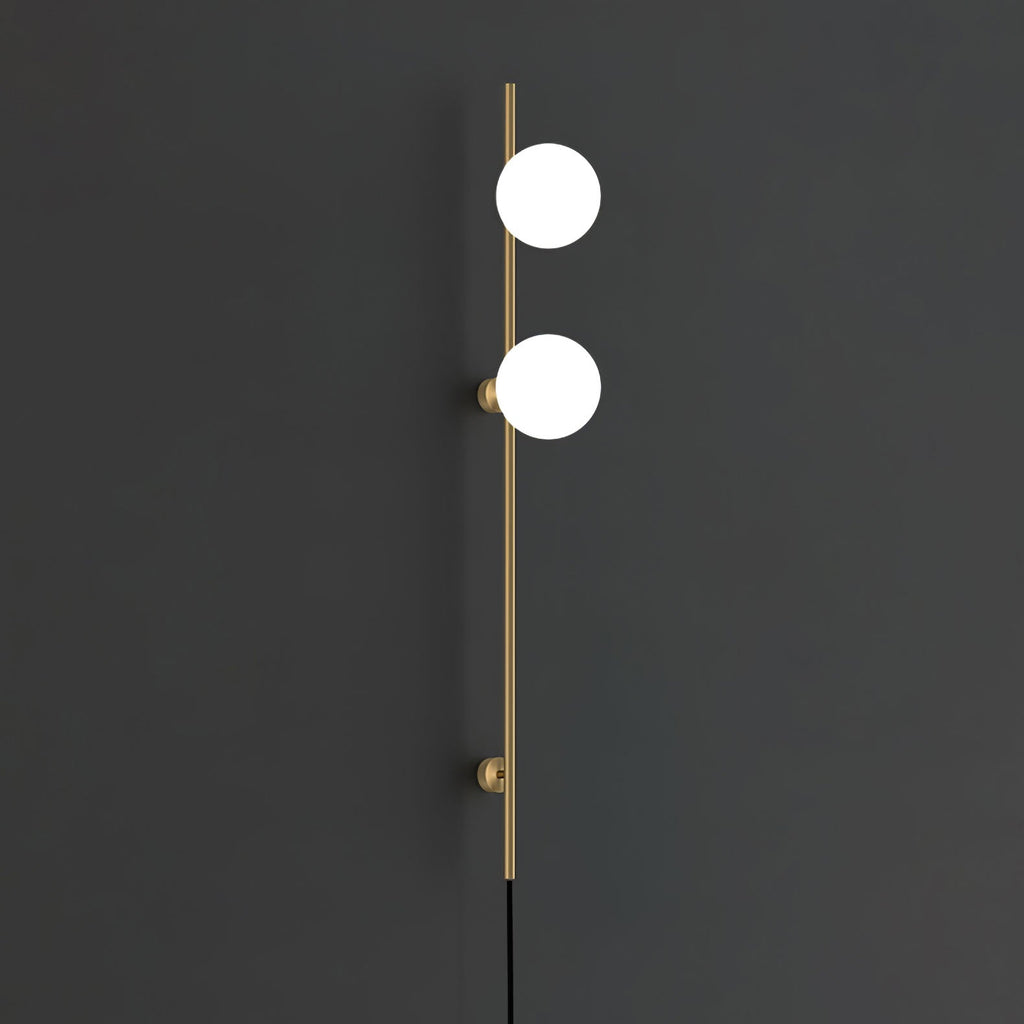 A Clérachic wall light in gold with two bright white globes on a slim rod and two round mounts, against a charcoal wall.