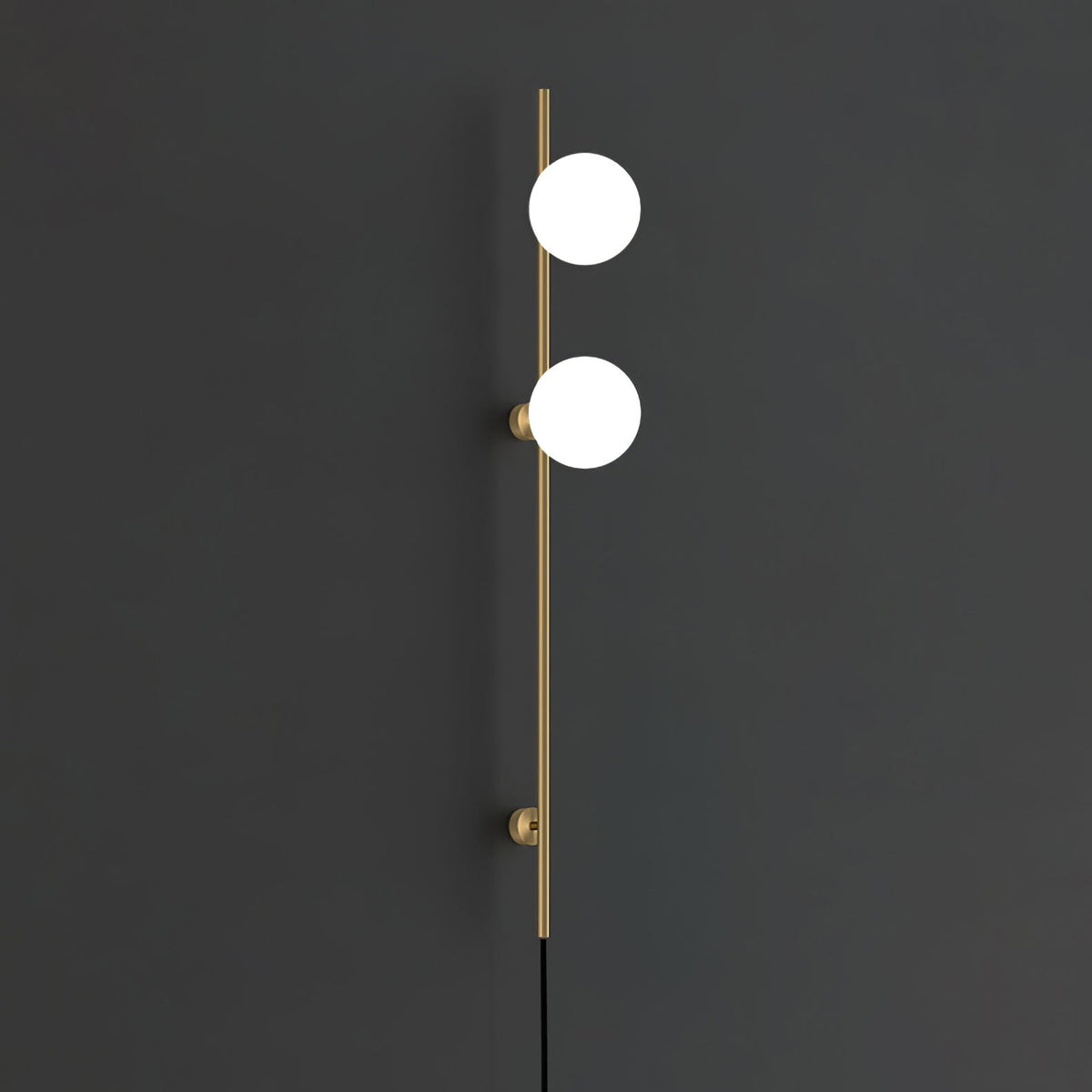 A Clérachic wall light in gold with two bright white globes on a slim rod and two round mounts, against a charcoal wall.