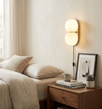 A lit white two-globe Clérachic wall sconce beside a bed, with a black cord running down to a walnut nightstand and framed print.