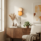 Clérachic two-globe wall sconce glowing in white and orange-pink, above a walnut sideboard with dried grasses and a boucle armchair.