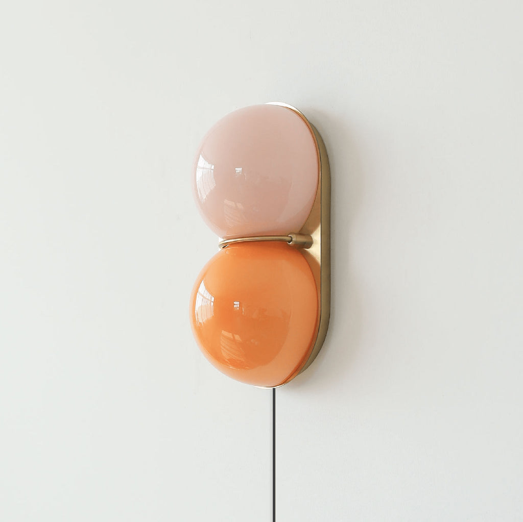 Clérachic two-globe wall sconce in pink and orange glass on a brass backplate, with a black cord hanging down a pale wall.