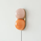 Clérachic two-globe wall sconce in pink and orange glass on a brass backplate, with a black cord hanging down a pale wall.