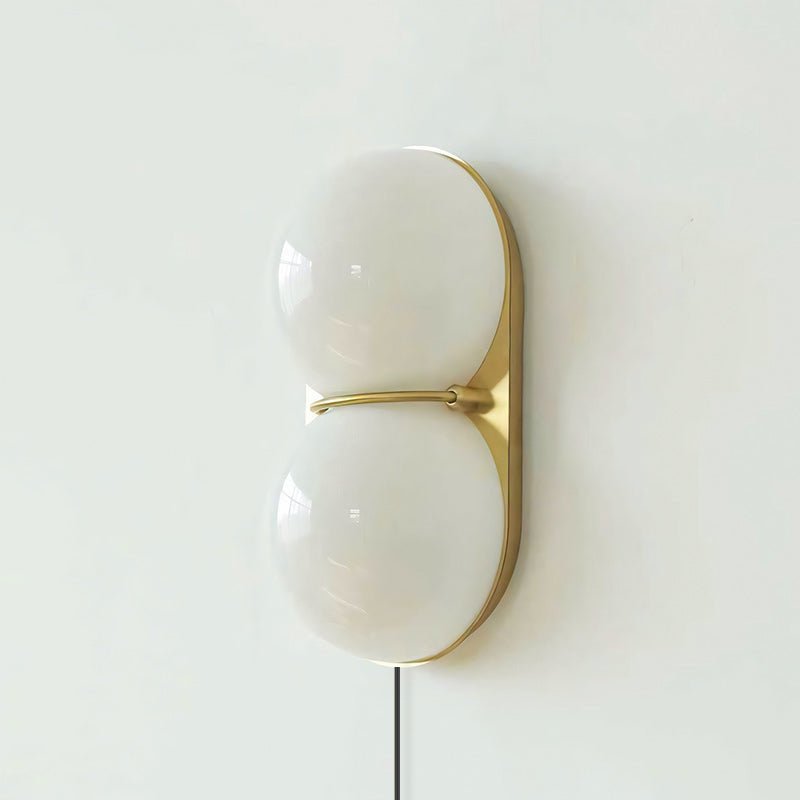 Close, front-on view of a white two-globe Clérachic wall sconce on a brass backplate, joined by a slim brass band.