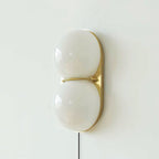 Close, front-on view of a white two-globe Clérachic wall sconce on a brass backplate, joined by a slim brass band.