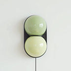 Clérachic wall sconce with two pale green globes inside a black oval backplate, with a black cord hanging below.