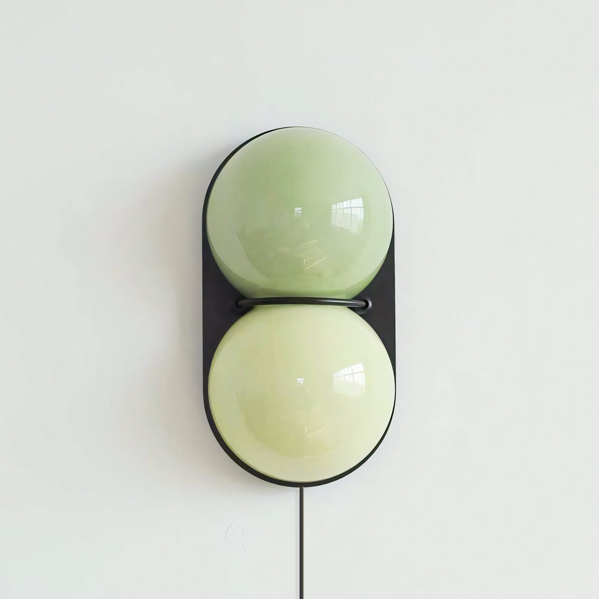 Clérachic wall sconce with two pale green globes inside a black oval backplate, with a black cord hanging below.